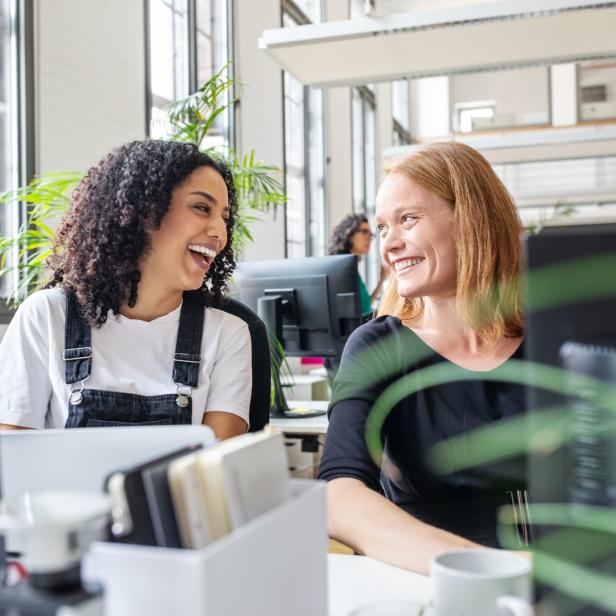 Zwei Frauen sitzen lachend an ihren Schreibtischen in einem Büro.