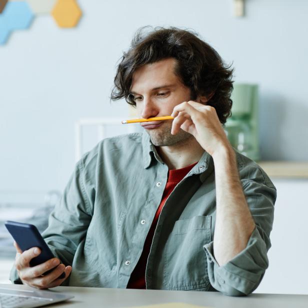 Ein Mann sitzt mit einem Stift im Mund vor einem Laptop und schaut auf sein Handy.