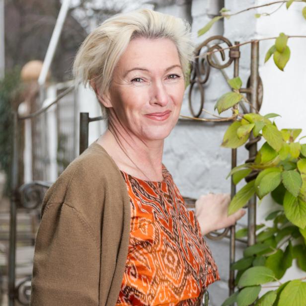 Eine Frau mit blonden Haaren lächelt neben einer begrünten Mauer.