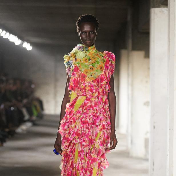 Ein Model präsentiert ein asymmetrisches, mehrlagiges Kleid mit Blumenmuster auf einem Laufsteg.