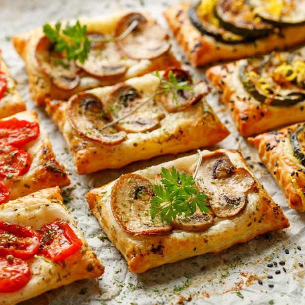 Blätterteig-Häppchen belegt mit Tomaten, Pilzen und Zucchini auf einem Backpapier.