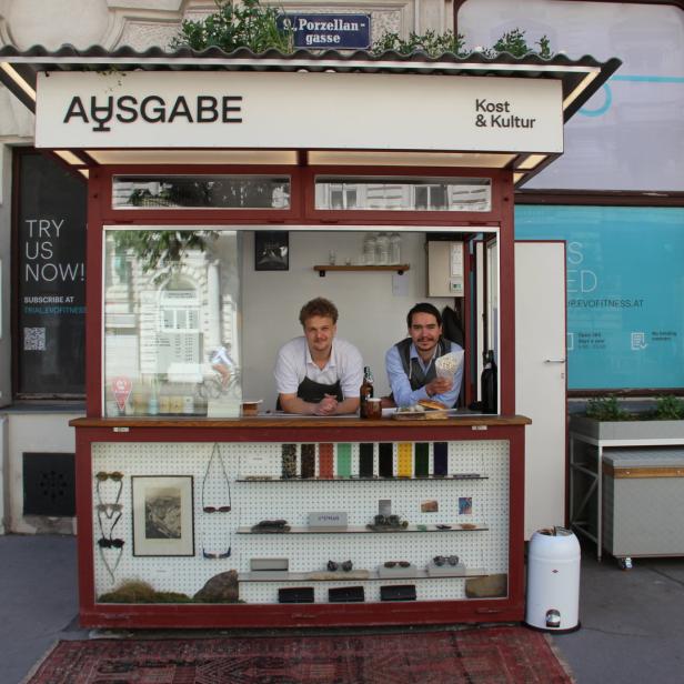 Zwei Männer stehen in einem Verkaufsstand mit der Aufschrift „Ausgabe – Kost & Kultur“.