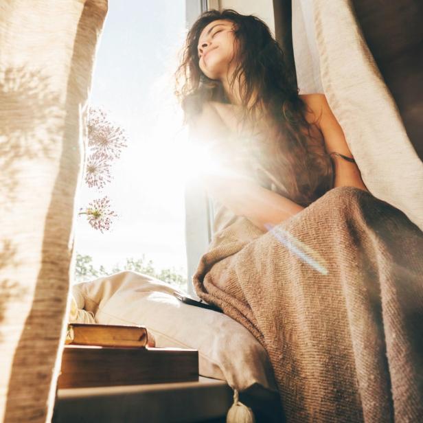 Eine Frau sitzt in einem Fenster, in eine Decke gehüllt, und genießt die Sonne.