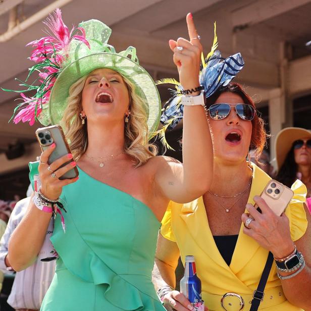 Zwei Frauen mit auffälligen Hüten jubeln bei einem Pferderennen.