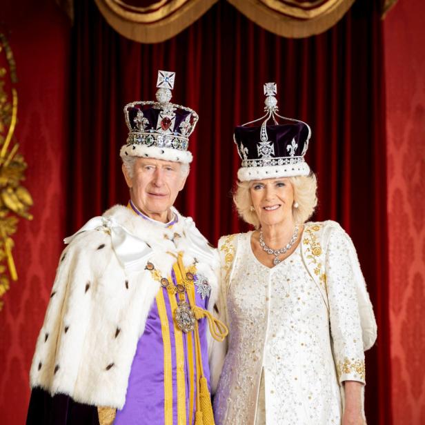 König Charles III. und Königin Camilla tragen Kronen und formelle Kleidung vor einem roten Vorhang.