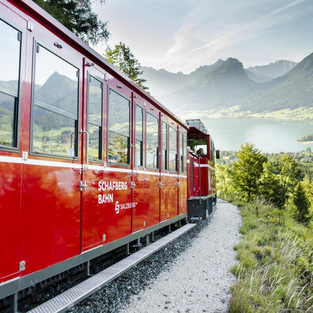 Die rote Schafbergbahn fährt vor einer Bergkulisse und einem See entlang.
