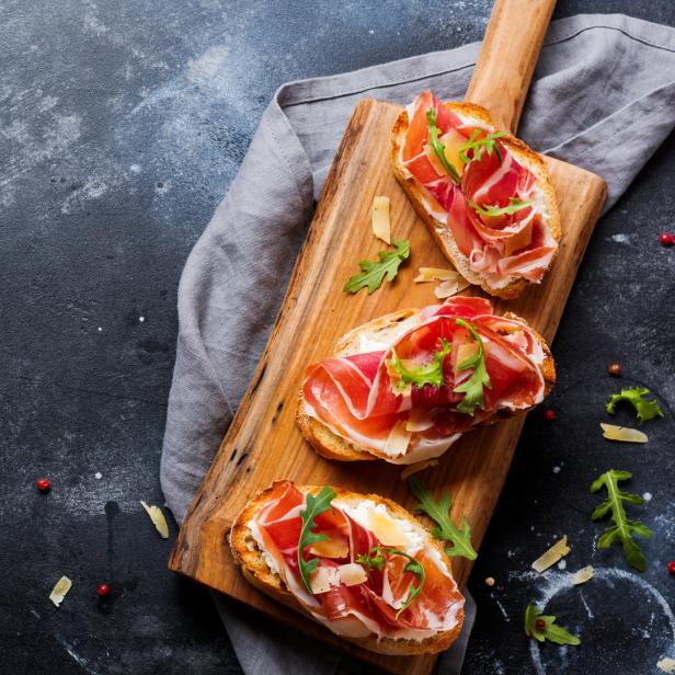 Drei Bruschetta mit Schinken, Käse und Rucola auf einem Holzbrett.