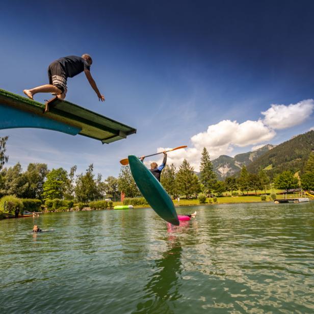 Ein Mann springt von einem Sprungturm in einen See, während eine Person im Kajak wartet.