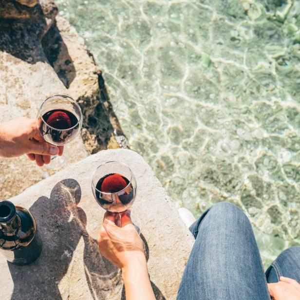 Zwei Personen stoßen mit Rotwein an, mit Blick auf das türkisfarbene Meer.
