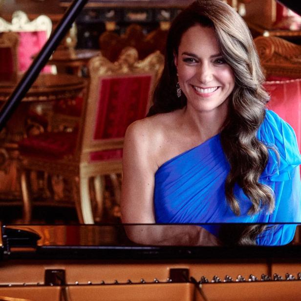 Kate Middleton lächelt in einem blauen Kleid an einem Klavier sitzend.
