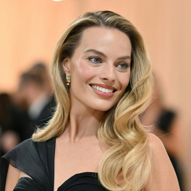 Margot Robbie lächelt in einem schwarzen Kleid mit goldenen Kettenakzenten.