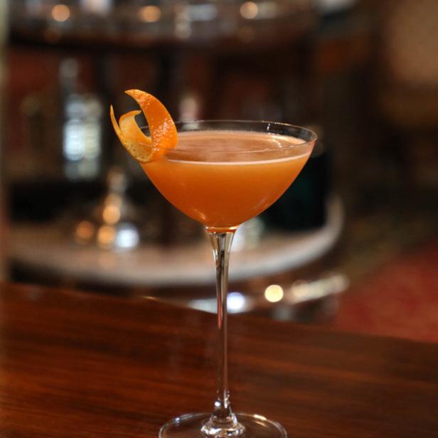 Ein Cocktail in einem Martiniglas, garniert mit einer Orangenschale, steht auf einer Bar.