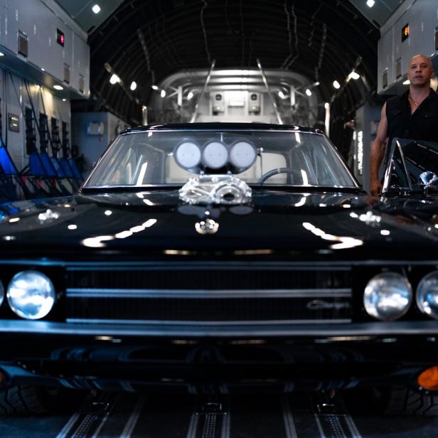 Vin Diesel posiert mit einem schwarzen Dodge Charger in einem Flugzeug.