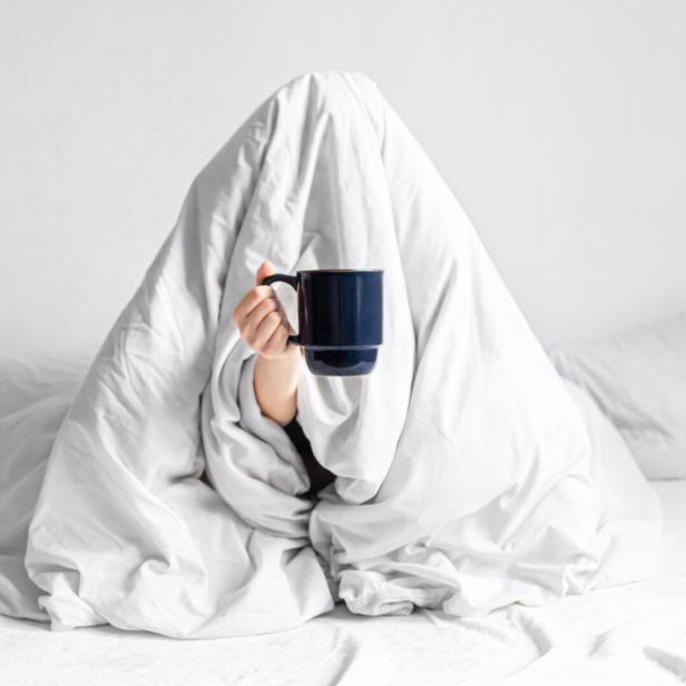 Eine junge Frau sitzt mit Kaffeetasse im Bett - die Decke über den Kopf gezogen.