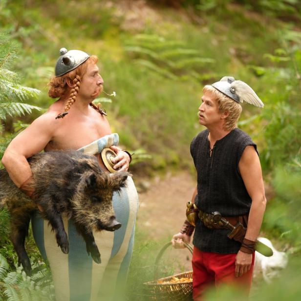 Asterix trägt ein Wildschwein, während Obelix mit einem Korb Pilze daneben steht.