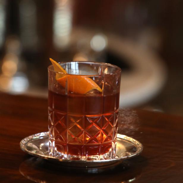 Ein Old Fashioned Cocktail mit Orangenschale auf einem silbernen Untersetzer.