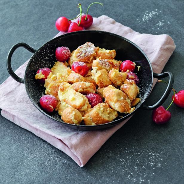 Kaiserschmarrn mit Kirschen in einer schwarzen Pfanne auf einem rosa Tuch angerichtet.