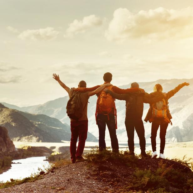 Freunde stehen mit ausgestreckten Armen auf einem Gipfel und blicken auf die Berglandschaft.