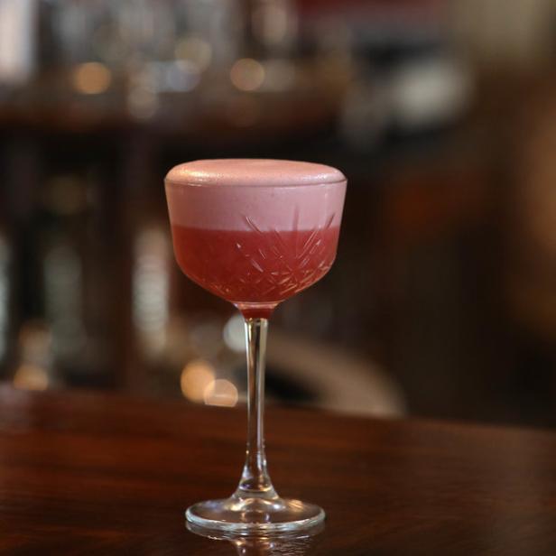 Ein rosa Cocktail mit Schaum steht auf einem dunklen Holztisch.
