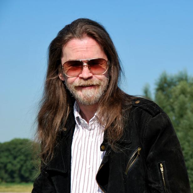 Ein Mann mit langen Haaren, Bart und Sonnenbrille vor blauem Himmel und Bäumen.