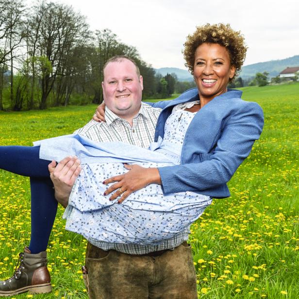 Arabella Kiesbauer wird von einem Mann auf einer Wiese getragen.