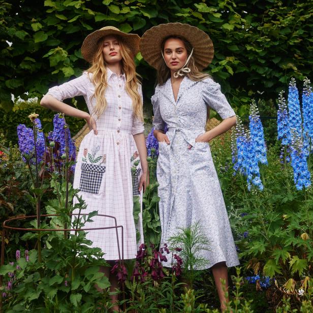 Zwei Frauen mit Strohhüten und Sommerkleidern stehen in einem blühenden Garten.
