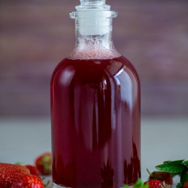 Eine Karaffe mit Erdbeersirup steht umgeben von frischen Erdbeeren.