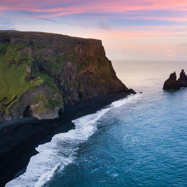 Blick auf Reynisfjara, Island, mit schwarzem Sandstrand, Basaltsäulen und Felsnadeln im Meer.