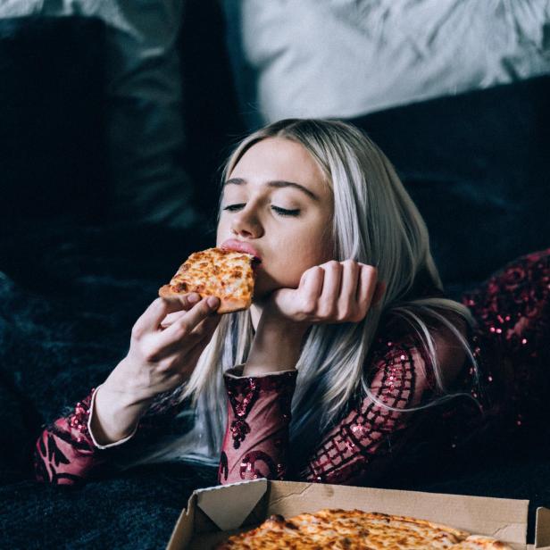 Eine Frau mit blonden Haaren isst ein Stück Pizza auf dem Bett.