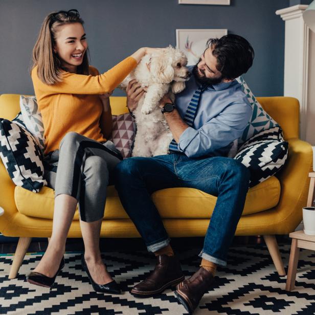 Ein Paar sitzt mit einem kleinen Hund auf einem gelben Sofa.