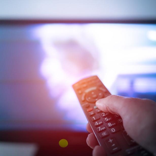 Eine Hand hält eine Fernbedienung vor einem eingeschalteten Fernseher.