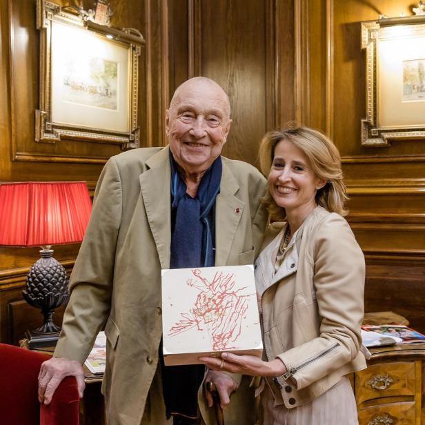 Georg Baselitz und Alexandra Winkler posieren mit einer Schachtel in einem holzgetäfelten Raum.