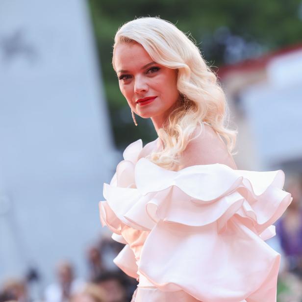 Eine Frau mit blonden, gewellten Haaren und einem rosa Kleid mit Rüschen.