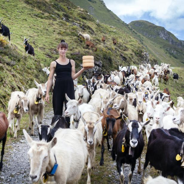 Eine Frau steht inmitten einer Ziegenherde in den Bergen und hält Käse.