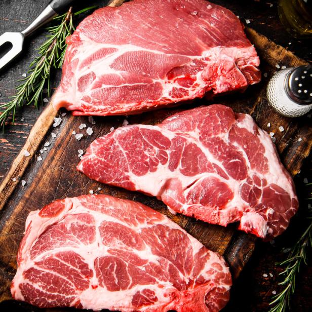 Drei rohe Schweinenackensteaks liegen auf einem Holzbrett mit Rosmarin und Gewürzen.