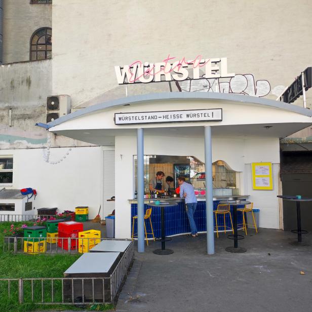 Ein Wurststand mit dem Schriftzug „Würstel“ und Gästen an der Theke.