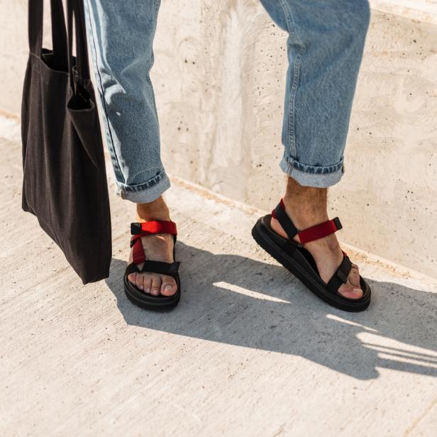 Eine Person in Jeans und Sandalen mit rotem Riemen steht neben einer schwarzen Stofftasche.