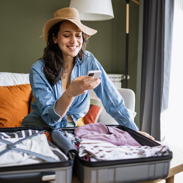 Eine lächelnde Frau mit Hut packt Kleidung in einen offenen Koffer und schaut auf ihr Smartphone.