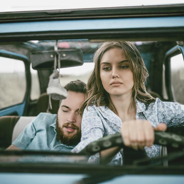 Eine junge Frau fährt einen blauen Oldtimer, während ein Mann auf dem Beifahrersitz schläft.
