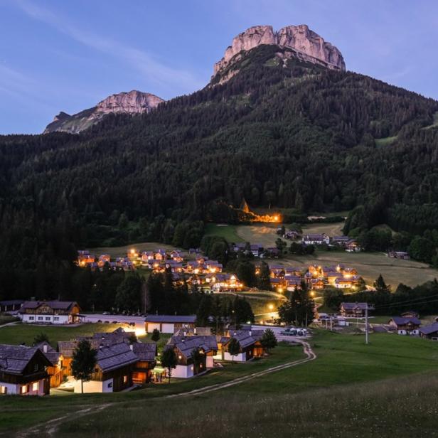 Ein Bergdorf liegt eingebettet in einer grünen Landschaft unter einem hohen Bergmassiv.