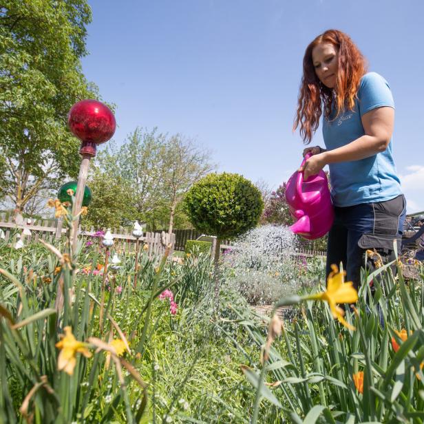 Eine Frau mit rotem Haar gießt Blumen in einem Garten mit einer pinkfarbenen Gießkanne.