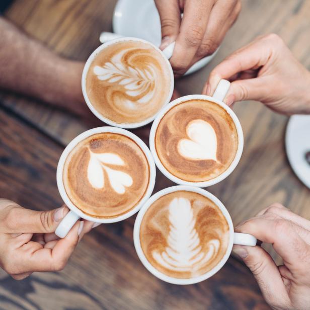 Vier Personen halten Tassen mit Latte Art auf einem Holztisch.