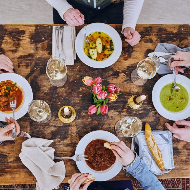 Ein Esstisch mit mehreren Personen, Tellern mit verschiedenen Gerichten, Wein und einem Blumenstrauß.