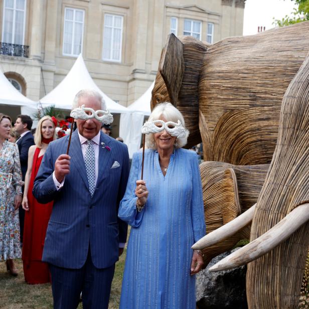 König Charles und Königin Camilla mit Masken vor einer Elefantenskulptur.