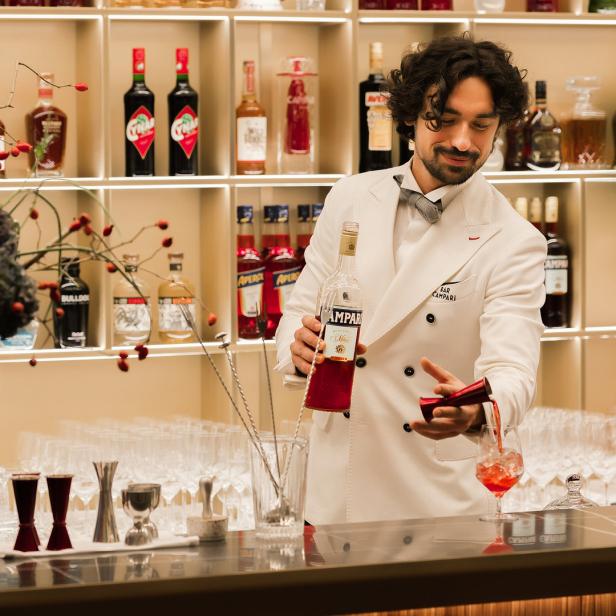 Ein Barkeeper in weißer Jacke mixt einen Cocktail mit Campari an einer Bar.