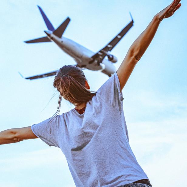 Eine Frau steht mit ausgestreckten Armen unter einem startenden Flugzeug.