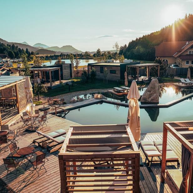 Blick auf ein Resort mit Schwimmteich, Holzhütten und Bergen im Hintergrund.