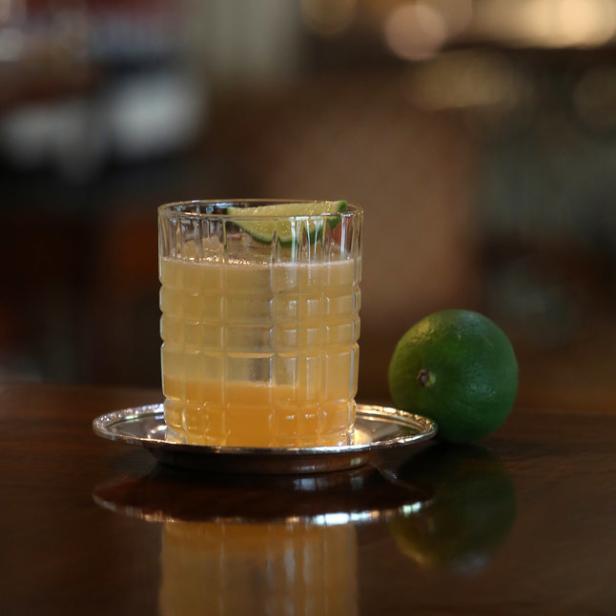 Ein Cocktail mit Limettenscheibe steht auf einem silbernen Untersetzer neben einer Limette.