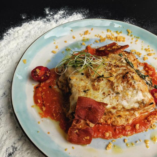 Eine Portion Lasagne auf einem Teller mit Tomatensauce und Speck garniert.