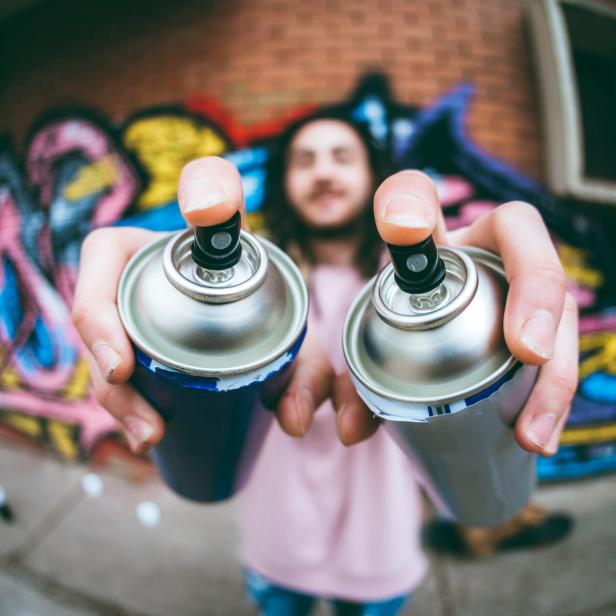 Eine Person hält zwei Sprühdosen vor einer mit Graffiti bemalten Wand.